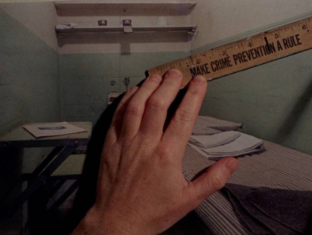 A screengrab from Malic Amalya's Living Lessons in the Museum of Order (2023), which features a large white -- probably male -- left hand holding a ruler, measuring the dimensions of a two-dimensional photo depiction of a prisoner's cell