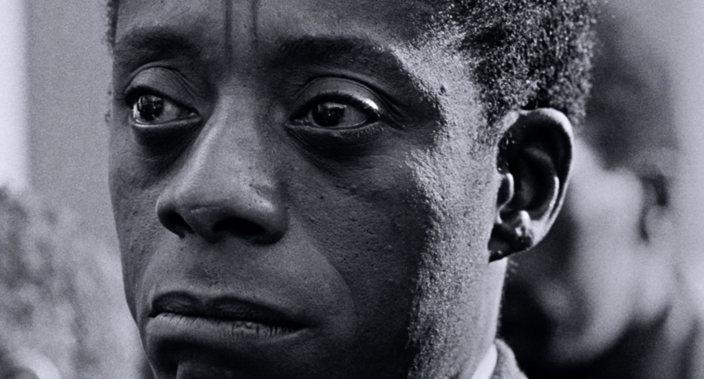 A tight close-up of James Baldwin's face -- he is slightly off center in the frame, and looking off-screen to the left, his eyes shining as if he is moved, his mouth downturned