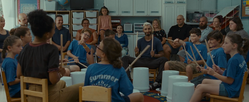 A still from Sound of Metal. A South Asian man with bleached blond hair, a dark beard and a blue T-shirt is sitting on a stool. His hands are raised, holding two sticks perpendicular to the ground, one in each hand. There is a white bucket face down on the ground in front of him. He is smiling, close-mouthed. To his right and left, middle-school aged children in oversized blue T-shirts are sitting on stools. They are also holding their sticks up, and have identical white buckets face down in front of them. They are sitting in a circle, so that we see the back of some of their heads. To either side of the man, a child is using their stick to point to a woman sitting at the back of the circle, with her back to the camera. She is Black, her tightly curled black hair is tied in a bun, and she is wearing a dark T-shirt. She is also holding two sticks perpendicular to the ground. Behind the circle of children, is a semi-circle of adults. Behind them are white cabinets and cubbies with white boxes inside. On top of one of the cabinets, a young white girl in a pink blouse and a half-up half-down hairstyle is sitting. She is looking intently at the circle of children.