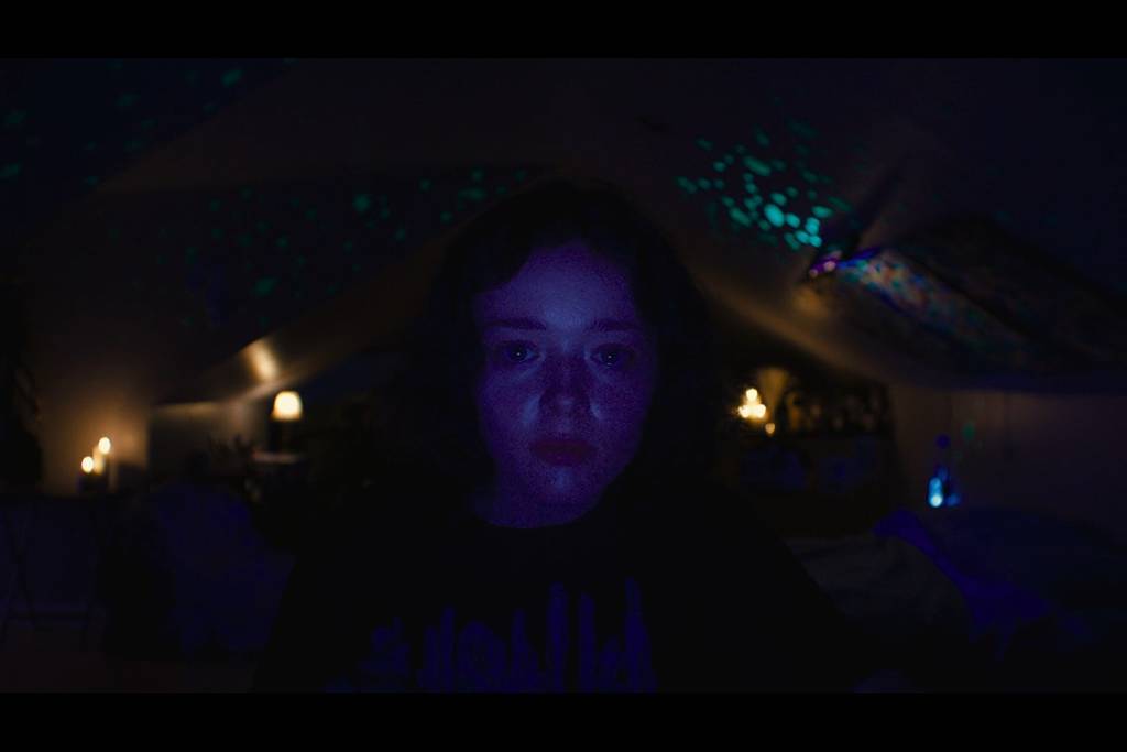 Casey’s face is illuminated by her computer screen as she stares into her webcam. There are many colors, primarily purple.
