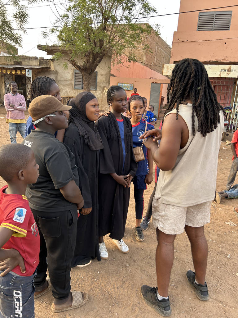 Straight-on color long shot of Madou Elijah Diarrassouba, his back to the camera, as he speaks to a group of young people of color outside.