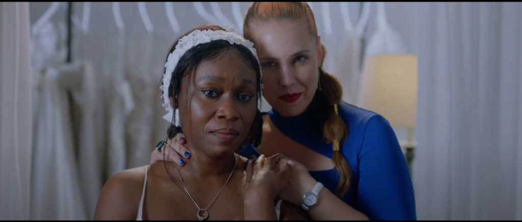 Straight-on color medium close-up of a Black woman and a white woman facing the camera, the white woman standing behind the Black woman, who wears a wedding dress.