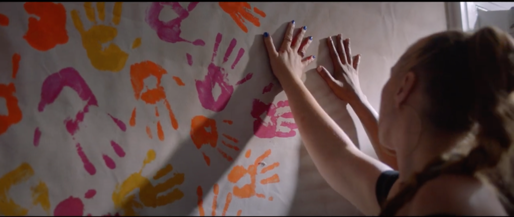 Low-angle color medium close-up of a white woman and a Black woman holding their hands up against a surface, making handprints; their backs are to the camera.