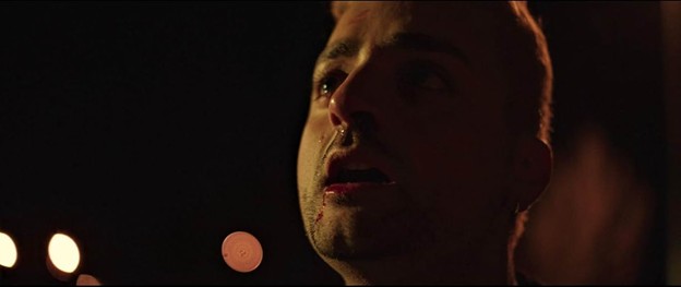 Straight-on color close-up of a person who appears male staring up and off camera to the left; lighting is harsh on the right side of his face as he presumably stands outside at night.
