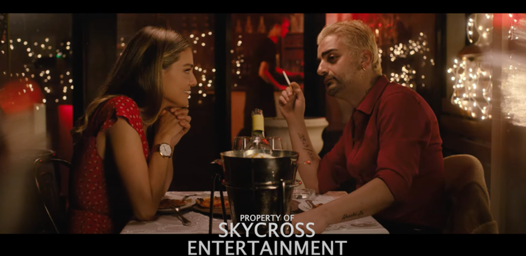 Straight-on color medium shot of a man and woman, dressed up for the occasion, sitting in profile at a table in a dark restaurant; fairy lights dominate the background; the man holds a cigarette in his right hand, looking slightly away from the woman, while the woman rests her chin on her clasped hands, gazing at the man.