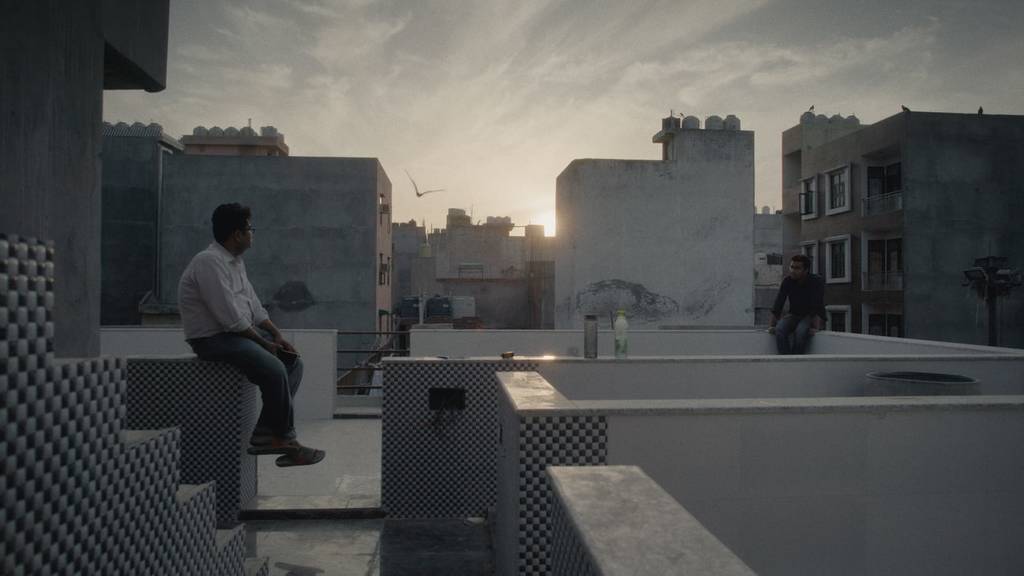 A straight-on color long shot of Nadeem Shehzad and Mohammad Saud sitting atop the wall that surrounds the roof of their building; birds fly around them in the sky as the sun sets.