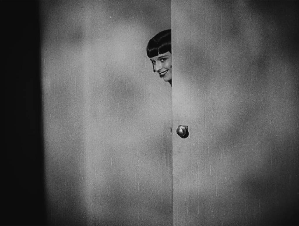 A straight-on black-and-white medium shot of Louise Brooks peering out from behind a door; her body is obscured by the door; only the left side of her face is revealed, in profile
