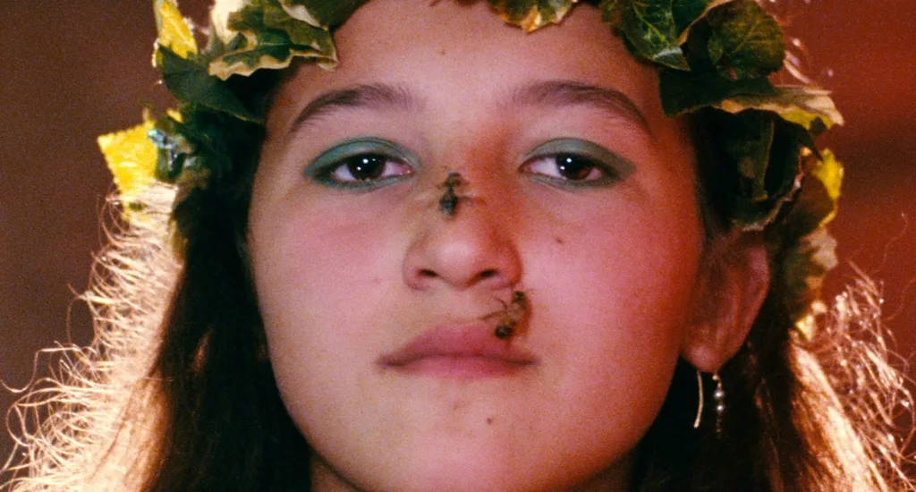 A straight-on color close-up of the face of a young girl, who appears white with long brown hair, staring directing at the camera while centered in the frame; she wears an ivy crown on her head, heavy green eyeshadow on her eyes, and gold dangly earrings; two bees are climbing on and around her nose.