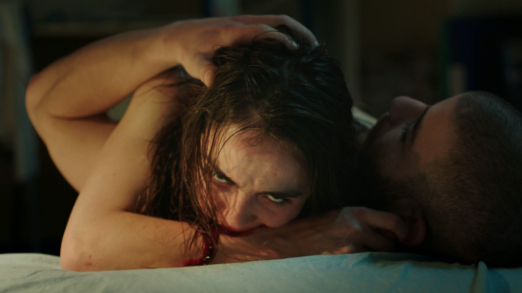 A straight-on color medium close-up, a side view, of two people, appearing to be a white man and a white woman, lying down on a bed; the woman is on top of the man, her head toward the camera as she bites her right arm, drawing blood; the man’s hand rests on top of her head.