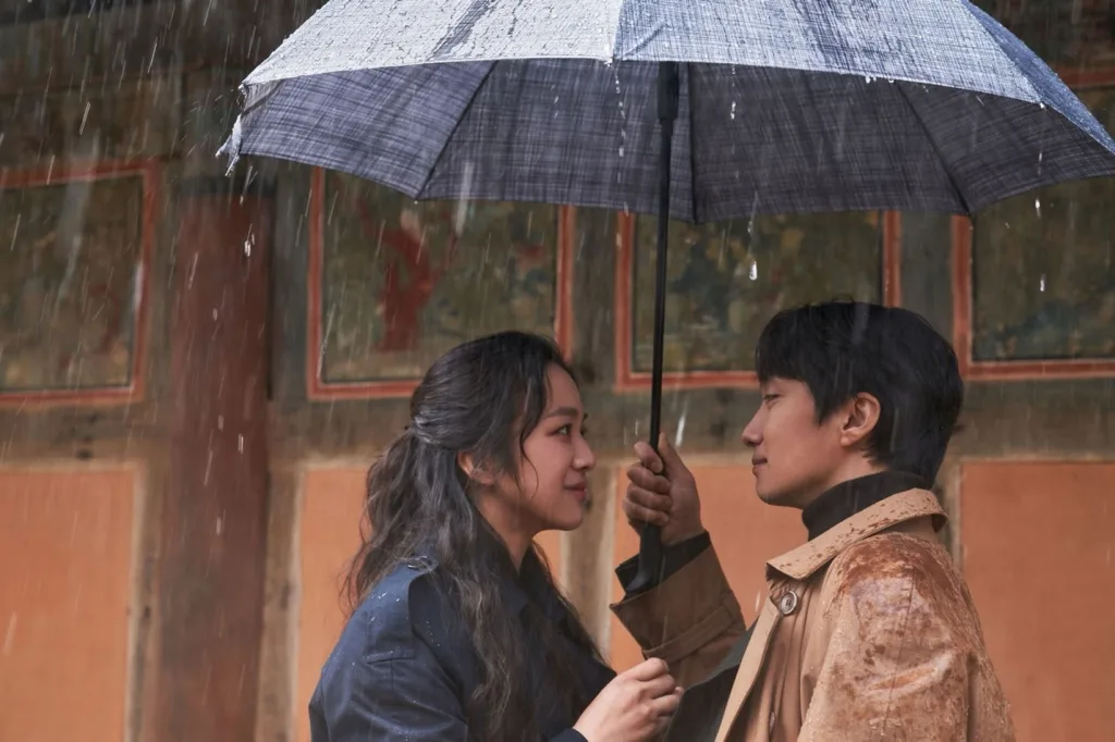 A young Asian woman with long black hair smiles into the face of a young Asian man with short black hair as they stand under an umbrella in the rain.