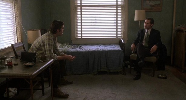 A straight-on color long shot of Matt Dillon and James Remar sitting perpendicularly in the interior of a bedroom, daylight visible through the blinds of the windows