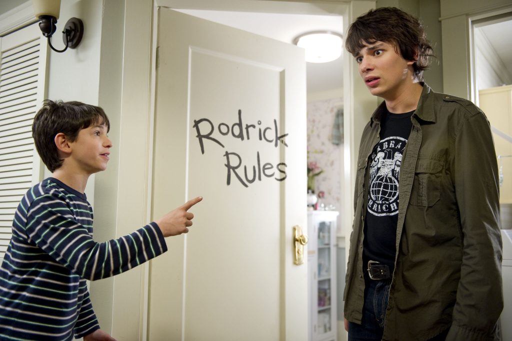 Two kids standing in a hallway in front of a bathroom door that has "Rodrick Rules" scrawled on it; the younger kid on the left is pointing at the older kid on the right
