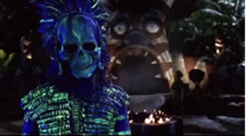 A scare character at Spooky Island stands outside at nighttime
The character wears a skull mask and tribal-style costume both painted with florescent blue and green paint, standing in front of a campfire and facade shaped to look like a face with a gaping mouth